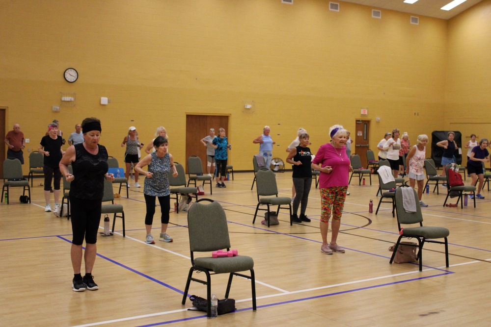 a gymnasium full of people in rows, standing next to chairs. There are water bottles and weights around